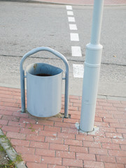 Fototapeta premium Metal urn near the road. A metal trash can is standing near a pillar on the sidewalk. Forging of garbage cans stands near a bench, on the sidewalk