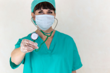 Blurred female doctor or nurse wearing face mask, hair net and green scrubs. Focus on stethoscope and hand with latex glove. Making eye contact. Plain pale background with copy space.