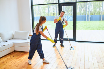 Young beautiful man and woman in blue uniform holding mops for washing living room, effective work, speaking during mopping floor, working