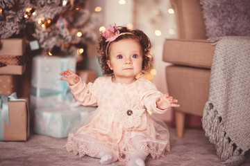 Beautiful baby girl princess 1 year old wearing stylish dress sitting on floor under Christmas tree closeup. Happy New Year. Childhood.