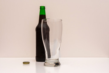 Empty transparent glass and an open green bottle with dark beer on a light background