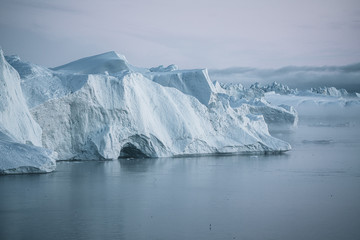 Fototapeta na wymiar Stranded icebergs in the fog at the mouth of the Icefjord near Ilulissat. Nature and landscapes of Greenland. Travel on the vessel among ices. Phenomenon of global warming. Coast in the sunset.
