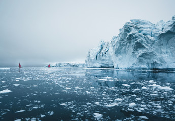 Beautiful red sailboat in the arctic next to a massive iceberg showing the scale. Cruising among...
