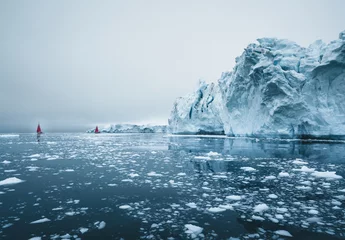 Wunderschönes rotes Segelboot in der Arktis neben einem massiven Eisberg, der die Skala zeigt. Kreuzfahrt zwischen schwimmenden Eisbergen im Gletscher der Diskobucht während der Mitternachtssonnensaison des Polarsommers Ilulissat, Diskobucht © Mathias