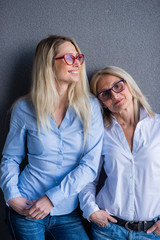 Portrait of two beautiful women of different generations against a gray wall. Family resemblance. Mother and daughter in a jeans shirt and glasses. Well-groomed elderly woman.