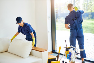 Two janitors in blue working uniform clean up roon with panoramic window, wearing yellow rubber...