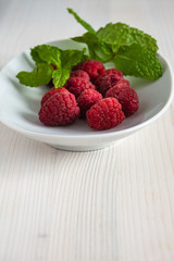 Raspberries with mint leaves on white plate and white wood in vertical with copy space