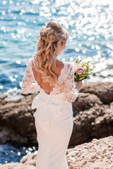 Beautiful bride in luxury wedding dress with bouquet at the sea side. Wedding by the sea. Bride walking around the sea near the place of the wedding ceremony. Back view