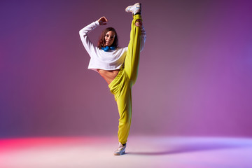 Flexible young dancer dressed in brightly coloured clothes, raising leg up, doing split leap in the air, work out of professional dancer, studio background