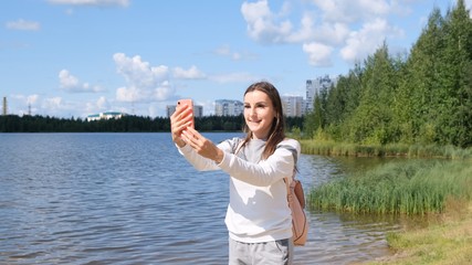 Beautiful girl with a backpack takes a selfie near the lake and talks on a cell phone on Skype.
