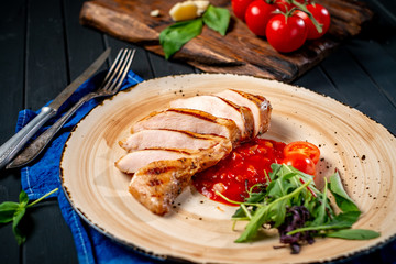 Fried sliced chicken fillet on a plate with tomatoes and arugula on a black wooden background. Diet dinner