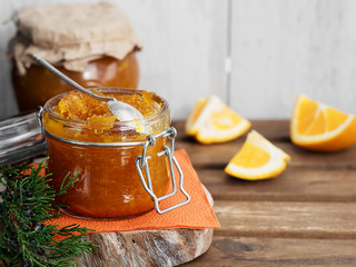 Tasty, homemade orange jam in a glass jar on a brown wooden background. Nearby there is a coniferous tree branch and orange slices. Dessert spoon in a jar of jam. Healthy citrus dessert. Close-up.