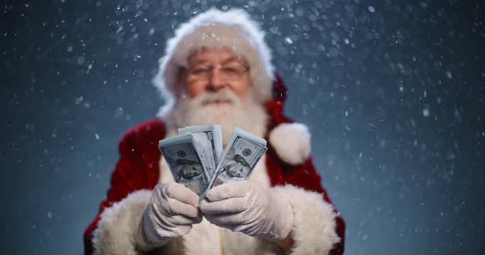 Santa clause looking at camera and showing packs of money in his hands, isolated on blue snowy background - christmas spirit concept close up 4k footage