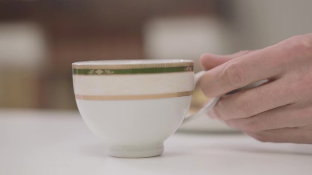 Close-up of white cup with tea or coffee standing at the table. Adult male Caucasian hand taking it and putting back in few moments.