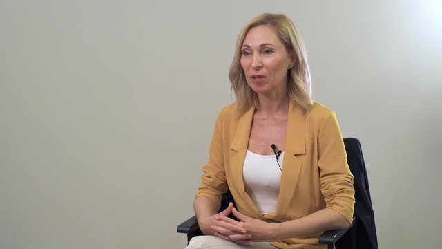Portrait of attractive blond Caucasian woman with grey eyes talking at the camera. Beautiful senior lady sitting at the chair and giving interview.