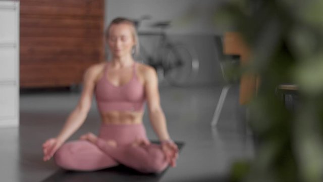 Young blonde woman makes yoga workout in her home loft.focus changes from the first plane plant to the woman in lotus pose