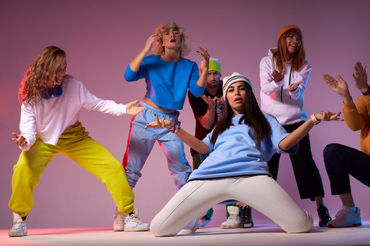 Beautiful Young Woman With Long Brunette Hair, In White Ca And Pants, Light Blue T Shirt, Standing On Knees, Spreading Hands Sideways With Confident Expression, Performing In The Middle Of Studio