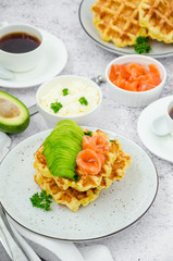 Cheese crispy waffles with cream cheese, smoked salmon and avocado for breakfast with a cup of coffee on a light stone background.
