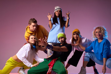 Team of professional dancers are taken photo in photo studio, dressed in brightly coloured sportswear, charming girl in white cap pointing fingers up demonstrating win gesture after dance performance