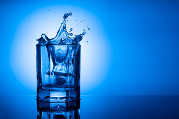 A splash of clear water in a glass tumbler from the fall of a piece of ice on a blue background. Mirror image. Horizontal Studio photography. copy space