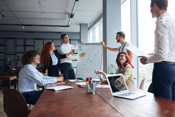 Young colleagues of man standing near flipchart explaining business project listen attentively to him. Business coworking