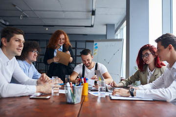 Young colleagues, caucasian business partners working as designers sit together at table in modern office with panoramic window. Papers, colorful pencils on table, active discussion