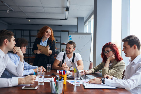 Personable business colleagues wearing formal wear gathered to discuss business project on table. Flipchart behind, laptops, pencils on table.