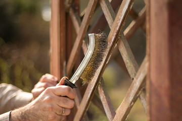 Prace remontowe w ogrodzie. Mężczyzna czyści deski ogrodowej pergoli drucianą szczotką. Usuwanie starej farby. - obrazy, fototapety, plakaty