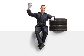 Auto mechanic sitting on a panel with tires and a wrench and waving at the camera - obrazy, fototapety, plakaty