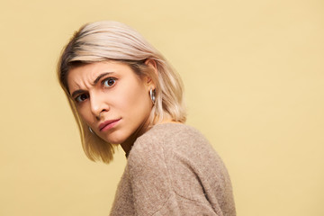 Studio shot of blonde young woman turning head to camera frowning not being in mood to talk, feeling disturbed, having indignant facial expression, posing isolated, wearing beige warm sweater