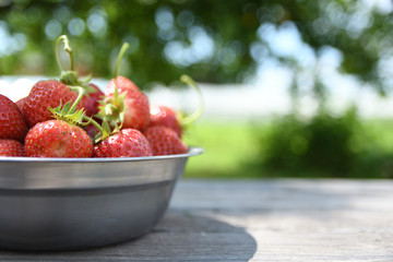  Strawberries in a bowl on a wooden table, outdoors on a green blurred background of leaves and trees