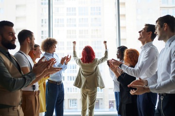 Young business leaders celebrate win of their colleague standing in the center in pose of winner with arms up, panoramic window background