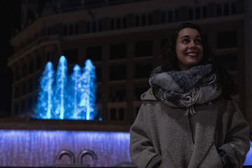 Retrato de una mujer de noche sonriendo con una bufanda delante de una fuente de agua
