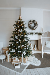 Christmas tree with boxes of gifts and a chair stands near the fireplace and other holiday decorations in white room