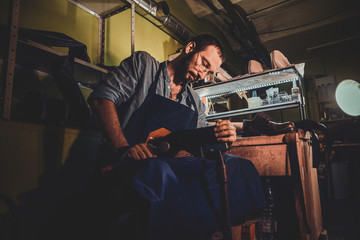 Young cobbler in glasses is working on shoe sole at his dark workplace.