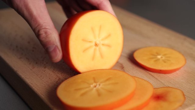 Chef's hand slices ripe orange persimmon on a wooden cutting board. The use of exotic tropical fruits in diets