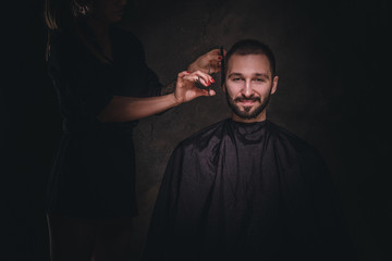 Dark portrait of young gentleman at hairdressing studio, he gets hair trimming from woman.