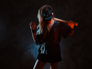 Lawbreaker female in spooky mask is standing at dark studio with bat in her hands. - obrazy, fototapety, plakaty