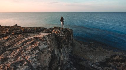 Fototapeta premium Woman traveler standing near sea on top of cliff in summer mountains and enjoying view of sea and nature. Cape Greco, Cyprus, Mediterranean Sea. Sunrise