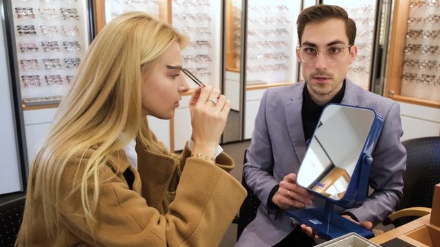 Elegant optical store consultant is giving a pair of glasses to the blond lady to try. He is holding a mirror. Lady is trying glasses and smile.