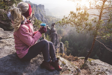 Female blond hiker outdoor clothing with coffee cup in hands on limestone rock enjoying sunset back...