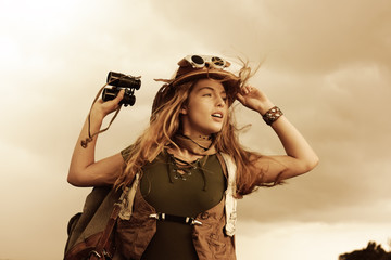 A young girl dresses up as an explorer. She wears a jungle hat and holds a binocular in her hand. The  wind passes through her hair as she observes the  area. - obrazy, fototapety, plakaty