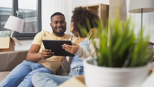 mortgage, people and real estate concept - happy african american couple with tablet pc computer sitting on sofa and kissing at new home