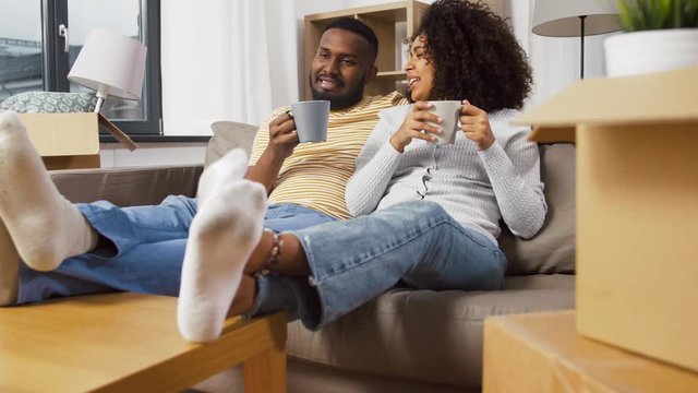 moving, people, repair and real estate concept - happy african american couple sitting on sofa and drinking coffee at new home