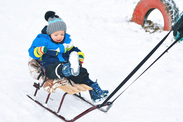 A little boy in a warm hat and a blue jacket sledding in the snow in winter.