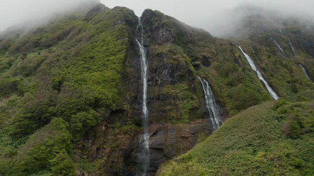 Waterfall in mountains. Strem of water on rocks among greenery. Aerial of cloudy Flores, Azores