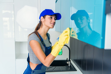 Cute young woman in workwear and protective rubber yellow gloves cleaning the furniture in modern hi-tech kitchen.