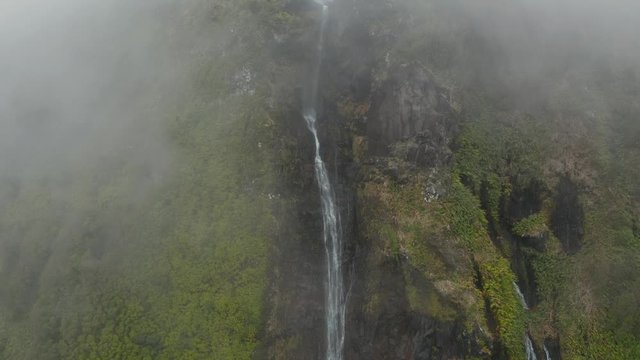 Aerial from cloud on high waterfall. Steep slopes, rocks and green plants. Aerial of Ferreiro, Flores, Azores