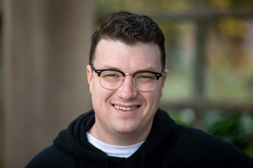 A young caucasian male wearing glasses, a white t-shirt and a black hoodie smiles and looks happy...