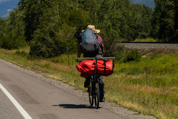 Mantraveling on bicycle is pictured from the back, while loaded with backpack and bike racks for his long journey. Natural environment in the background.